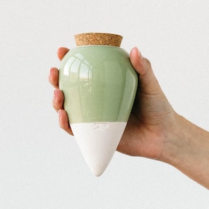 Może przedstawiać: Dwukolorowy ceramiczny wazon z jasnozieloną górną częścią i białą dolną częścią. Wazon ma korek i zaokrąglony korpus, który zwęża się ku dołowi. Wazon jest trzymany w dłoni.
