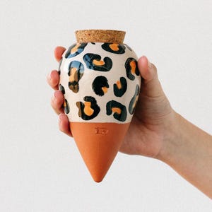 Könnte beinhalten: Eine Keramikvase mit cremefarbenem Körper, verziert mit schwarzen und orangefarbenen Leopardenflecken. Die Vase hat einen konisch geformten Sockel in Terrakottafarbe und einen Korken. Die Vase wird von einer Person gehalten.