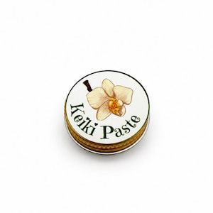 May include: A small, round, metallic container with a screw-top lid. The lid features a white background with a detailed illustration of a cream-colored orchid flower and the dark green text Keiki Paste printed in a curved, decorative font.