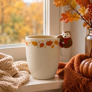 Peut inclure: Un mug en céramique crème orné d'une bande d'illustrations de feuilles d'automne orange et jaunes sur le bord. Une figurine d'ours brun en relief portant une écharpe rouge est fixée sur le côté du mug et sert de poignée décorative.
