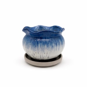 May include: A small, round ceramic planter with a wavy rim, glazed in a gradient of blue and white. The planter sits on a matching gray saucer. This decorative pot is ideal for small plants and succulents.