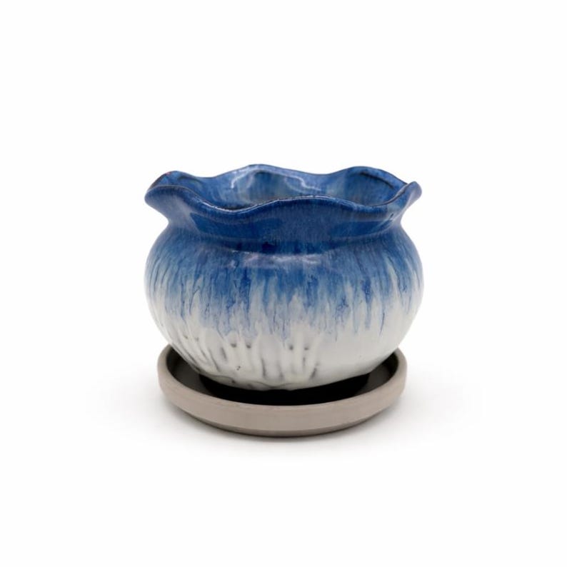 May include: A small, round ceramic planter with a wavy rim, glazed in a gradient of blue and white. The planter sits on a matching gray saucer. This decorative pot is ideal for small plants and succulents.
