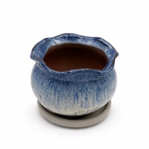 May include: A small, round ceramic planter with a wavy rim, glazed in shades of blue and white. The interior is brown, and it sits on a matching gray saucer. This decorative pot is ideal for small plants and succulents.
