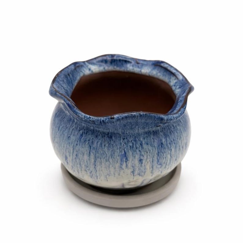 May include: A small, round ceramic planter with a wavy rim, glazed in shades of blue and white. The interior is brown, and it sits on a matching gray saucer. This decorative pot is ideal for small plants and succulents.