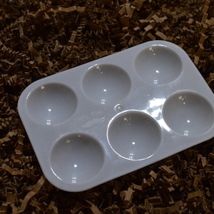 Puede incluir: Una paleta de pintura blanca rectangular con seis pozos circulares. La paleta es de plástico brillante y descansa sobre un lecho de papel triturado marrón. Diseñada para mezclar pinturas.