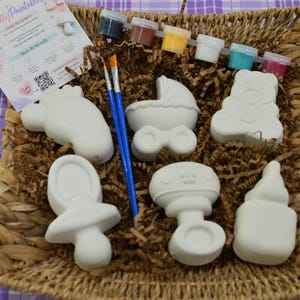 Puede incluir: Un kit de pintura para bebés con seis formas de cerámica, incluyendo un chupete, un biberón y un osito de peluche. El kit incluye botes de pintura, pinceles y una tarjeta con instrucciones, todo en una cesta de mimbre.