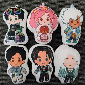 May include: A collection of six plush dolls with cartoon character designs. Each doll features a unique character with distinct outfits and hairstyles. The dolls are primarily white with colorful accents, including rainbow wheels on one doll's wheelchair.