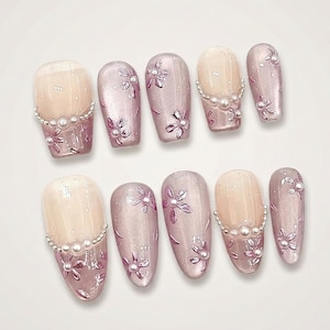 Op de afbeelding: Een set van tien kunstnagels. De nagels hebben een lichtroze en nude kleurenschema met bloemmotieven en parelaccenten. De nagels zijn ovaal van vorm en gerangschikt op een wit oppervlak.