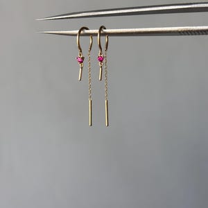 Op de afbeelding: Een paar goudkleurige oorhangers. Elke oorbel heeft een kleine, ronde, rode edelsteen, een gouden staaf en een fijne gouden ketting. Ze worden gepresenteerd tegen een grijze achtergrond.