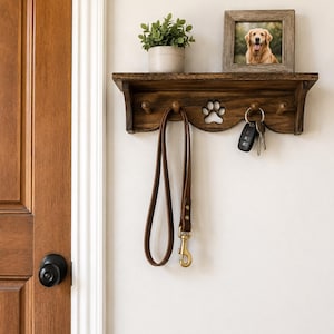 May include: A wooden wall-mounted shelf with a dog paw cutout, hooks, and a framed photo of a golden retriever. A brown leather dog leash hangs below. A small potted plant sits on the shelf.
