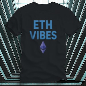 Op de afbeelding: Zwart t-shirt met de blauwe tekst "ETH VIBES" boven een paars Ethereum-symbool. Het ontwerp is gecentreerd op de voorkant van het shirt. Het shirt is gemaakt van een zacht materiaal.