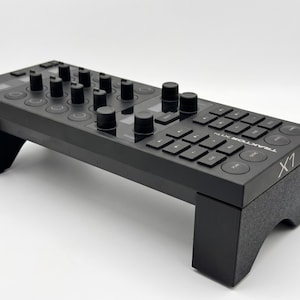 Può includere: Controller DJ nero con manopole, pulsanti e display. Il controller è etichettato "TRAKTOR X1 MK2" e "X1". Il dispositivo è sollevato su due supporti neri.
