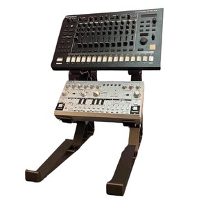 Può includere: Un supporto in plastica nera a due livelli che sostiene una drum machine Roland TR-8 nera sul ripiano superiore e un sintetizzatore Behringer TD-3 argentato su quello inferiore. Il supporto presenta una struttura robusta e il logo KREO3D.