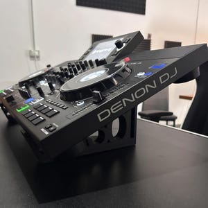 Puede incluir: Un controlador DJ autónomo Denon DJ Prime 4 de color negro sobre un soporte inclinado negro. El dispositivo cuenta con una pantalla táctil central, dos grandes jog wheels con anillos iluminados, pads de rendimiento y varios controles.