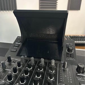 Può includere: Un controller DJ nero dotato di uno schermo centrale con un paraluce nero pieghevole contrassegnato dalla scritta OKREOBU LAB. Il dispositivo include numerose manopole rotanti, cursori e pulsanti neri per il mixaggio audio, con etichette bianche.