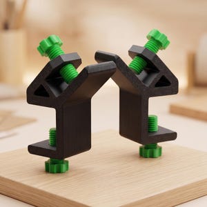Op de afbeelding: Twee zwarte C-klemmen met groene stelschroeven zijn gemonteerd op een lichtgekleurd houten oppervlak. De klemmen zijn ontworpen om materialen bij elkaar te houden. De groene schroeven zorgen voor de klemkracht.