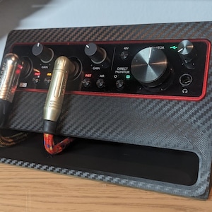 May include: A black and gray audio interface with a carbon fiber pattern. The interface has knobs, buttons, and input jacks. Two gold and black XLR cables are plugged into the front. The interface sits on a wooden surface.