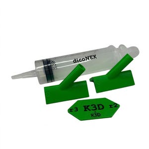 Può includere: Una siringa in plastica trasparente con tacche nere e la scritta "dicoNEX" stampata sul cilindro. Sono inclusi due ugelli in plastica verde angolati e una piastra esagonale in plastica verde con il testo nero "r3 K3D r2" e "K3D".