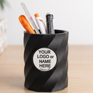 May include: A black cylindrical pencil holder with a spiralling textured surface. A circular white emblem on the front features the black text "YOUR LOGO or NAME HERE" and "KREO3D LAB". The holder contains an orange pen, a metal ruler, a white pen, and a black marker.
