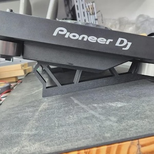 Può includere: Un supporto nero per controller Pioneer DJ con finitura opaca e una struttura di supporto geometrica a telaio aperto. Il supporto è progettato per sollevare l'attrezzatura, con due piedini circolari argentati visibili sotto il dispositivo.