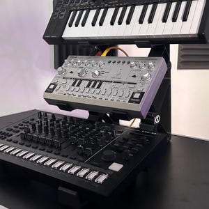 Może przedstawiać: Czarny, wielopoziomowy stojak na syntezatory, na którym znajduje się czarny kontroler MIDI Behringer Swing na górnej półce, srebrny syntezator basowy Behringer TD-3 na środkowej półce oraz czarny automat perkusyjny Roland TR-8 na podstawie.