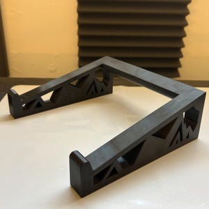Puede incluir: Un soporte negro para portátil con un diseño geométrico recortado en los paneles laterales. El soporte tiene dos brazos paralelos conectados por una barra transversal trasera, con bordes delanteros curvados hacia arriba para asegurar el dispositivo.