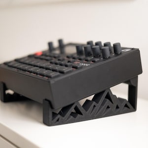 以下が含まれることがあります： 黒色の電子音楽制作デバイス。キーボードとノブがあり、黒色のスタンドに置かれています。スタンドは幾何学的な山のデザインです。デバイスは白い表面にあります。