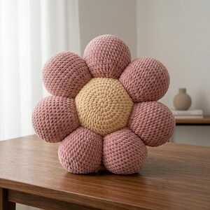 May include: A handmade, flower-shaped cushion with a light pink petal design and a tan center. The cushion is made of a soft, textured material and sits on a wooden table. The petals are round and evenly spaced.