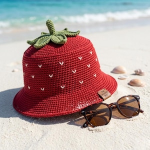 May include: A red crochet bucket hat shaped like a strawberry, with a green stem and leaves. White stitches form the strawberry seeds. The hat sits on a sandy beach next to sunglasses. The tag reads "THE CROCHET NEST".