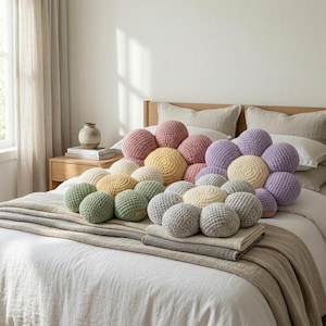 May include: A collection of colorful, round, crocheted pillows arranged on a bed. The pillows are in shades of pink, yellow, green, gray, and lavender. The bed is made with white linens and a neutral-toned blanket.
