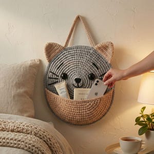 K&ouml;nnte beinhalten: Ein grau-beiger Wandorganizer in Katzenform aus H&auml;kelarbeit mit einer Tasche. Die Katze hat schwarze Augen, Schnurrhaare und Ohren. Die Tasche enth&auml;lt eine Creme, ein Buch und ein Smartphone. Eine Hand greift nach dem Telefon.