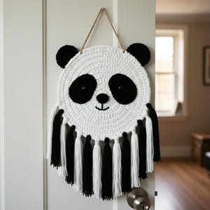 May include: A handmade, white crochet panda face wall hanging with black detailing. The panda features black eyes, ears, and a nose, with a smiling mouth. Black and white tassels hang from the bottom. The wall hanging is suspended by a brown string.