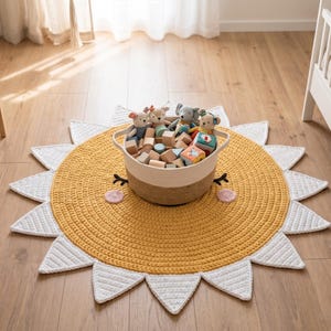 Puede incluir: Una alfombra amarilla y blanca con forma de sol y textura tejida. Una cesta tejida llena de bloques de madera y animales de peluche se encuentra en el centro. La alfombra tiene un dise&ntilde;o de cara sonriente con mejillas rosas y ojos negros.