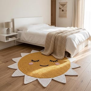 Puede incluir: Una alfombra de ganchillo amarilla y blanca con forma de sol sonriente con los ojos cerrados. La alfombra est&aacute; sobre un suelo de madera en un dormitorio, con una cama y una mesita de noche en el fondo.