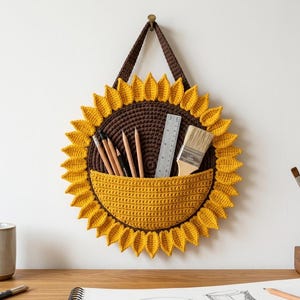 May include: A sunflower-shaped crochet wall organizer with a dark brown center and bright yellow petals. It features a semi-circular pocket holding several pencils, a clear plastic ruler, and a wooden paintbrush with light-colored bristles.