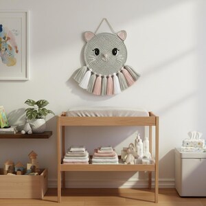 May include: A light wood changing table with a grey changing pad. A grey, pink, and white cat-shaped wall hanging is above the table. The table has a lower shelf with folded towels and baby supplies.