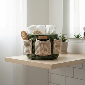 May include: A green crochet basket with three beige pockets, holding white towels, a wooden hairbrush, and toiletries. The basket sits on a light wood shelf, ideal for bathroom organization. A small leather tag is attached to the basket.
