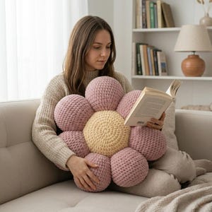 May include: A large, handmade flower-shaped cushion in shades of pink and yellow. The cushion is made of a soft, textured material and has a central yellow circle surrounded by pink petals. The cushion is being held by a person.