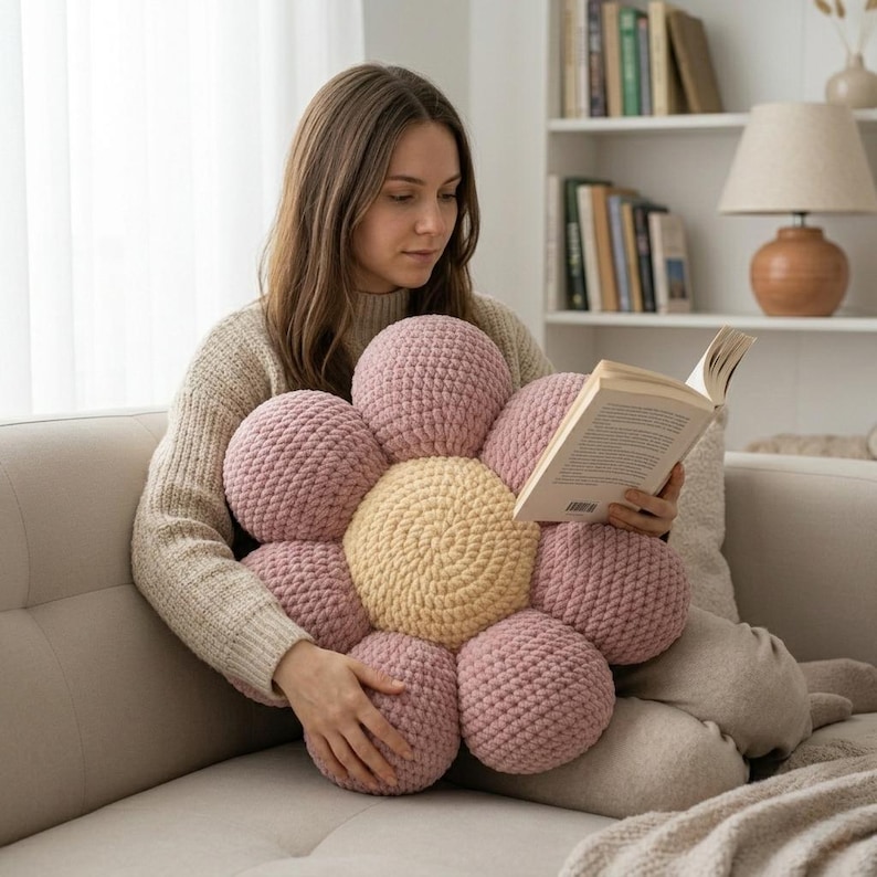 May include: A large, handmade flower-shaped cushion in shades of pink and yellow. The cushion is made of a soft, textured material and has a central yellow circle surrounded by pink petals. The cushion is being held by a person.