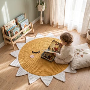 Può includere: Un tappeto a forma di sole giallo e bianco con un design a faccina sorridente. Il tappeto è realizzato in un materiale spesso e strutturato ed è circondato da raggi triangolari bianchi. Un bambino è seduto sul tappeto, che legge un libro.