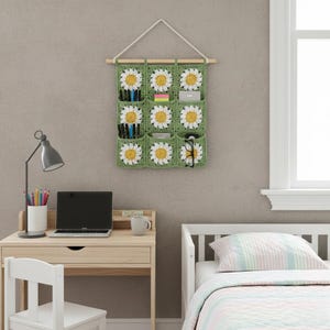 May include: A green crochet wall organizer with nine pockets, each adorned with a white and yellow daisy design. The organizer is suspended from a wooden dowel and holds pens, sticky notes, and glasses. It hangs on a neutral-colored wall.