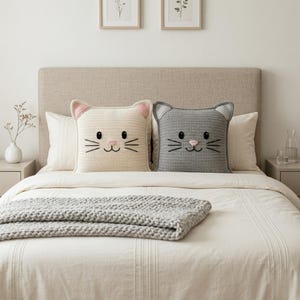 Peut inclure: Deux coussins en crochet en forme de chat, un cr&egrave;me et un gris, sont pos&eacute;s sur un lit. Les coussins pr&eacute;sentent des d&eacute;tails brod&eacute;s pour les yeux, le nez et les moustaches, avec des accents roses sur les oreilles et le nez. Une couverture grise en tricot est au pied du lit.