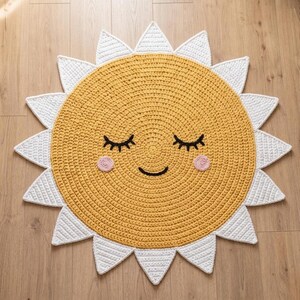 Puede incluir: Alfombra de ganchillo amarilla hecha a mano con forma de sol sonriente. El sol tiene rayos blancos, pesta&ntilde;as negras, una sonrisa negra y mejillas rosas. Un art&iacute;culo decorativo para una habitaci&oacute;n infantil.