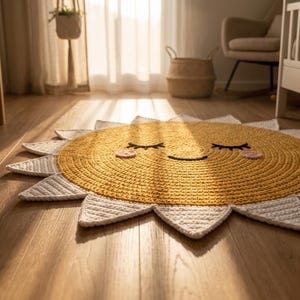 Puede incluir: Una alfombra con forma de sol tejida en crochet amarillo y blanco con una cara sonriente. La alfombra tiene un centro amarillo con pesta&ntilde;as negras y una boca sonriente. Los rayos del sol son blancos y puntiagudos. La alfombra est&aacute; sobre un suelo de madera.