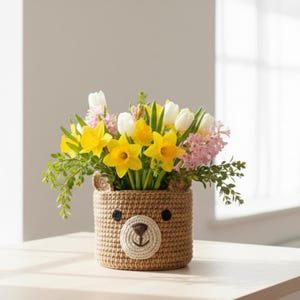 Può includere: Un vaso di fiori a forma di orso marrone all'uncinetto, pieno di narcisi gialli, tulipani bianchi e giacinti rosa. Il vaso ha una faccia cucita con occhi a bottone neri e un muso color crema.