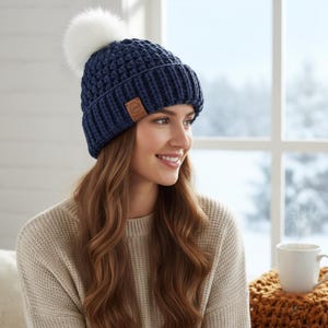 Puede incluir: Un gorro de punto azul marino con un pompón de piel sintética blanco. El gorro presenta un patrón texturizado y una pequeña etiqueta de cuero marrón. La mujer lleva un suéter beige.