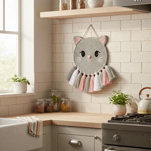 May include: A grey crochet cat wall hanging with pink inner ears, black eyes, and white whiskers. The cat's face is round with a pink nose. Below the cat's face are grey, pink, and white tassels. The wall hanging is displayed in a kitchen.