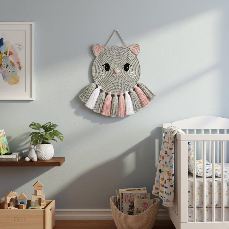 May include: A grey, crocheted cat wall hanging with pink ears and a pink nose. The cat has black eyes and white whiskers. Below the cat's face are tassels in grey, white, and pink. The wall hanging is displayed on a light blue wall.