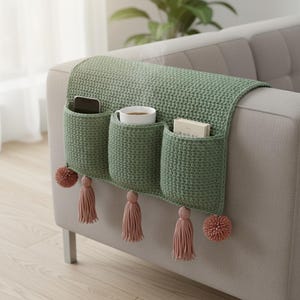 May include: A sage green crochet sofa arm organizer with three pockets, holding a phone, a mug, and a book. The organizer has pink tassels and pom-poms for decoration. The item is placed on a gray sofa.