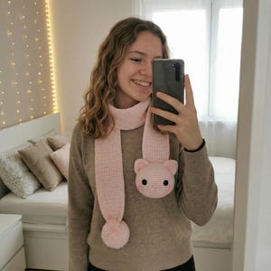 May include: A light pink crocheted scarf with a cat face design and a pom-pom. The scarf is made of soft yarn and features a cat head with black eyes and pink ears. The scarf is worn around the neck.
